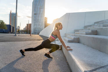 Side view of slim female athlete in sportswear stretching leg near staircase while exercising in back lit and looking forward - ADSF19869