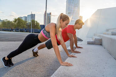 Ganzkörper-Seitenansicht eines entschlossenen jungen aktiven Mannes und einer Frau in Sportkleidung, die während eines gemeinsamen Fitness-Trainings auf einer städtischen Straße Liegestütze gegen Betonstufen machen - ADSF19870