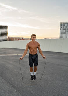 Young smiling muscular male athlete jumping rope on roadway during workout under shiny sky in evening and looking forward - ADSF19876