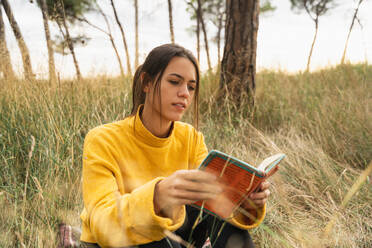 Sorglose Frau sitzt im Herbst in einem Feld und genießt eine interessante Geschichte in einem Buch - ADSF19878