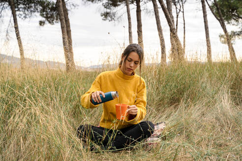 Gelassene Frau sitzt auf trockenem Gras einer Wiese und gießt ein heißes Getränk aus einer Thermoskanne in eine Tasse, während sie das Wochenende in der Natur genießt - ADSF19880