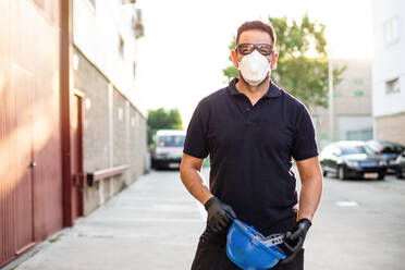 Erwachsener männlicher Mechaniker mit Schutzbrille und Atemschutzmaske, der einen blauen Schutzhelm in den Händen hält und in die Kamera schaut, während er in der Nähe eines Werkstattgebäudes auf der Straße steht - ADSF19894