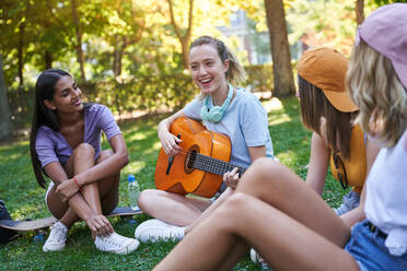 Gruppe von verschiedenen Teenager-Freundinnen spielen Gitarre und haben Spaß, während sie zusammen auf grünem Gras im Park sitzen und im Sommer chillen - ADSF19899