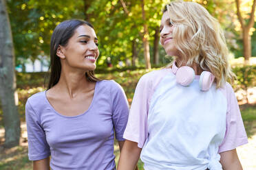 Happy young blond female with headphones on neck chatting with ethnic girlfriend while spending sunny summer day together and strolling in green park - ADSF19900