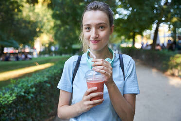 Joyful blonde teen female with cold drink spending summer day in park looking at camera - ADSF19905