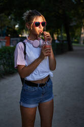 Hipster teen female with sunglasses enjoying cold milkshake while resting in park in summer day - ADSF19906