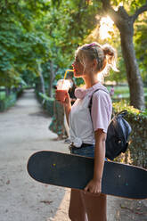 Seitenansicht von Hipster Teenager-Skaterin trägt Skateboard und genießt kalten Milchshake beim Ausruhen im Park im Sommer Tag - ADSF19908
