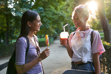 Joyful diverse teen female friends with fruit popsicle and cold drink chatting happily while spending summer day together in park - ADSF19909