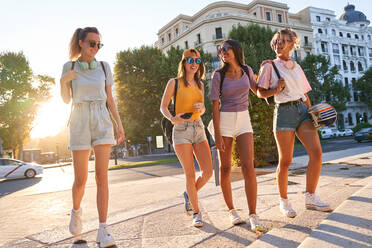 Gruppe fröhlicher multirassischer Teenager-Mädchen in Freizeitkleidung mit Kopfhörern und Skateboard, die Spaß haben und fröhlich plaudern, während sie im Sommer zusammen auf einer Stadtstraße spazieren gehen - ADSF19910