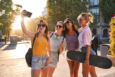 Fröhliche trendige junge multirassische Freundinnen mit Sonnenbrillen, Rucksäcken und Skateboard, die ein Selfie mit ihrem Handy machen, während sie sich auf einem städtischen Platz im Sommer versammeln - ADSF19911