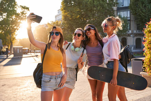 Fröhliche trendige junge multirassische Freundinnen mit Sonnenbrillen, Rucksäcken und Skateboard, die ein Selfie mit ihrem Handy machen, während sie sich auf einem städtischen Platz im Sommer versammeln - ADSF19911