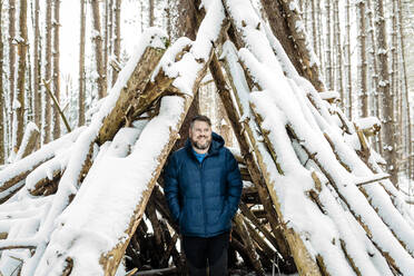 Lächelnder Mann steht mit den Händen in den Taschen in einem mit Schnee bedeckten Tipi im Wald - AWAF00031