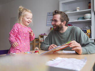 Happy father showing book to cute daughter on table at home - LAF02627