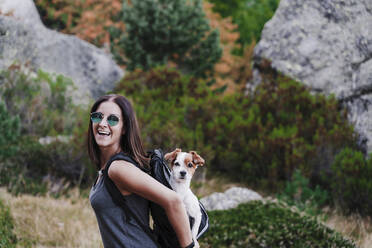 Cheerful woman carrying dog in backpack while hiking on mountain - EBBF02181