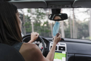 Nahaufnahme einer Frau, die ihre Gesichtsmaske an den Rückspiegel im Auto hängt - EBBF02190