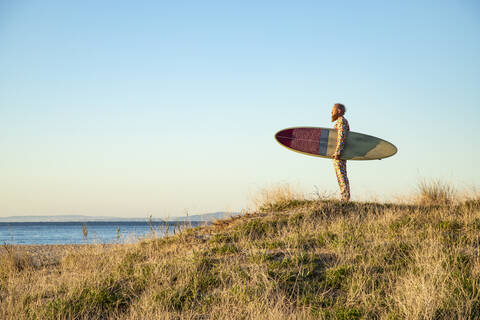 Mann schaut auf das Meer, während er mit einem Surfbrett am Strand steht, lizenzfreies Stockfoto