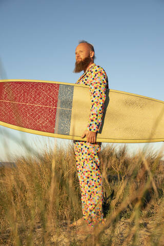 Mann in buntem Anzug mit Surfbrett schaut weg, während er gegen den Himmel steht, lizenzfreies Stockfoto