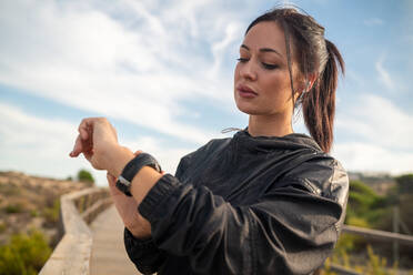 Active young brunette in black sports outfit standing on wooden pathway and checking data on fitness tracker after training in nature - ADSF19925