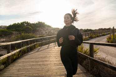 Aktive junge Frau in schwarzer Sportkleidung beim Joggen auf einem Holzweg während eines Outdoor-Fitnesstrainings in der Natur - ADSF19926