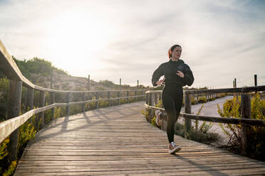 Aktive junge Frau in schwarzer Sportkleidung joggt auf einem Holzpfad während eines Fitnesstrainings in der Natur - ADSF19928