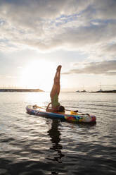 Side view full body of unrecognizable slim female in swimwear doing Supported Shoulder Stand pose while practicing yoga on floating paddle board at sunset time - ADSF19940