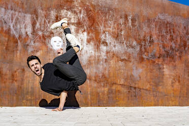 Young man with white mask shouting while doing handstand against brown wall - GGGF00803