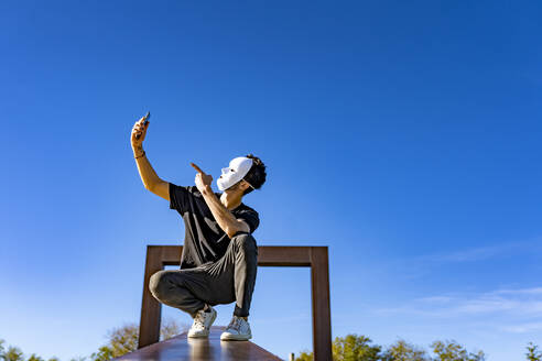 Man with white mask taking selfie through mobile phone while crouching against sky - GGGF00815