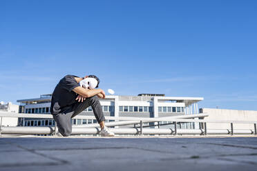 Man with white mask crouching on rooftop against sky - GGGF00821