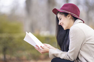 Lächelnde Frau mit Hut, die im Park sitzend ein Buch liest - GGGF00833