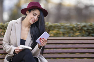 Beautiful woman wearing hat using mobile phone while sitting on bench in park - GGGF00834