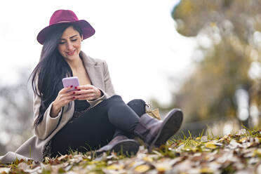 Lächelnde schöne Frau, die ein Mobiltelefon benutzt, während sie auf einem Grundstück gegen den klaren Himmel im Park sitzt - GGGF00835