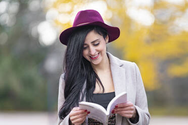 Lächelnde schöne Frau mit Hut, die am Wochenende im Park ein Buch liest - GGGF00838