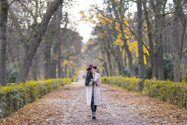 Mid adult woman reading book while walking on land amidst trees in park - GGGF00840