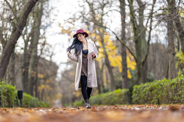 Smiling mid adult woman wearing hat walking on land in park during autumn - GGGF00842