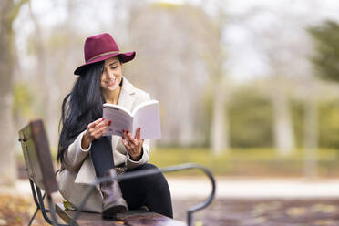 Schöne Frau mit langen Haaren, die ein Buch liest, während sie auf einer Bank im Park sitzt - GGGF00843