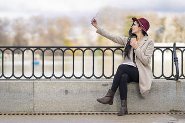 Mittlere erwachsene Frau, die einen Kaffee hält und ein Selfie mit ihrem Smartphone macht, während sie auf einer Stützmauer sitzt - GGGF00846