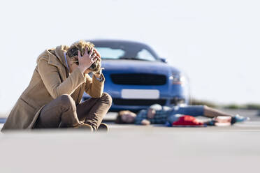 Verzweifelter junger Mann auf dem Boden sitzend mit einem Jungen im Hintergrund, der nach einem Unfall neben dem Auto liegt - GGGF00851