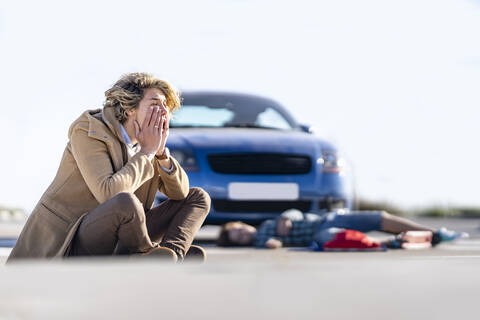 Desperate young man sitting on ground with boy in background lying by car after accident stock photo