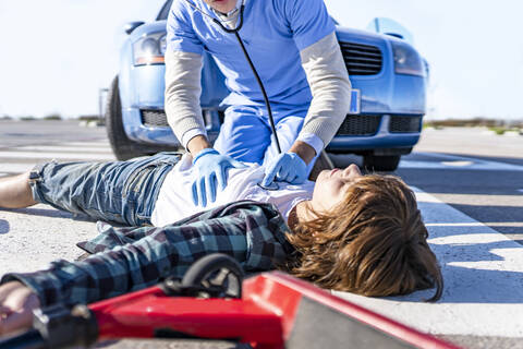 Male rescue worker with stethoscope examining boy lying on road after car accident stock photo