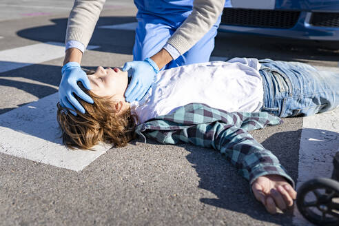 Doctor checking unconscious boy lying on road after car accident - GGGF00860