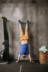 Upside down view of athletic man in protective mask performing handstand near wall in modern gym during workout - ADSF19976