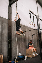 Low angle of muscular sportsman training in gym and climbing rope during functional workout - ADSF19989