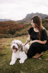 Cute fluffy Spanish Water Dog giving paw for smiling female owner while sitting together on grassy hill in mountains - ADSF19993