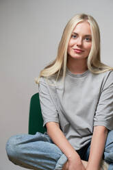 Young cool female in stylish outfit sitting on chair on gray background and looking at camera - ADSF20003