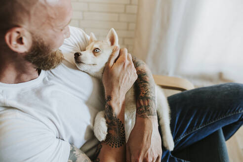 Mid adult man embracing dog while sitting on chair at home - MIMFF00411