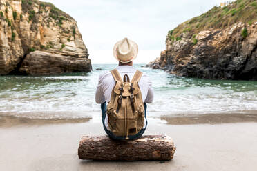 Rückenansicht eines nicht erkennbaren männlichen Rucksacktouristen mit Hut, der auf einem Holzklotz an der Küste nahe dem Meer mit felsigen Klippen sitzt und sich während einer Sommerreise entspannt - ADSF20027