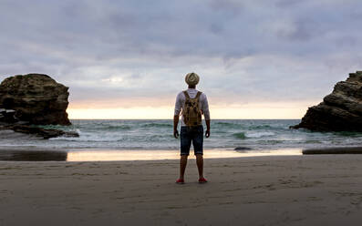 Full body back view of unrecognizable man with backpack standing on empty sandy shore against waving sea and cloudy sky and enjoying fresh breeze and freedom in summer evening during sunset - ADSF20028