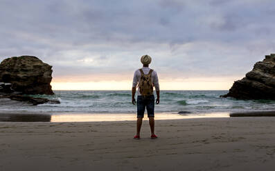 Ganzkörper Rückenansicht von unerkennbaren Mann mit Rucksack stehen auf leeren Sandstrand gegen wogenden Meer und bewölkten Himmel und genießen frische Brise und Freiheit im Sommer Abend während des Sonnenuntergangs - ADSF20028