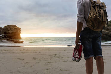 Rückenansicht des anonymen männlichen Rucksacktouristen mit Turnschuhen in der Hand, der allein am Sandstrand nahe dem Meer steht und den Sonnenuntergang bewundert, während er sich nach einer Wanderung in der Natur entspannt - ADSF20029