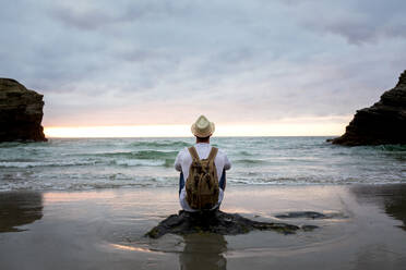 Back view of anonymous male traveler sitting on a rock at sandy coast enjoying views during summer vacation near sea at sunset - ADSF20032